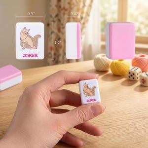 May include: White mahjong tile with a cartoon cat illustration and the word "JOKER". The tile is 0.9 inches wide and 1.2 inches tall. Other tiles are pink and white. The game pieces are on a wooden table.