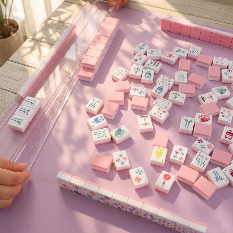 May include: A pink and white Mahjong set with tiles featuring various symbols and designs. The tiles are scattered on a light purple surface, with some arranged in rows. The set includes a clear plastic tile holder.