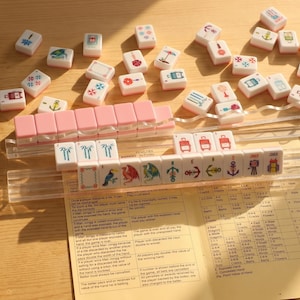 May include: A Mahjong set with pink and white tiles, featuring various symbols and illustrations. The tiles are arranged on a wooden surface, with a clear plastic rack and a rule sheet visible.