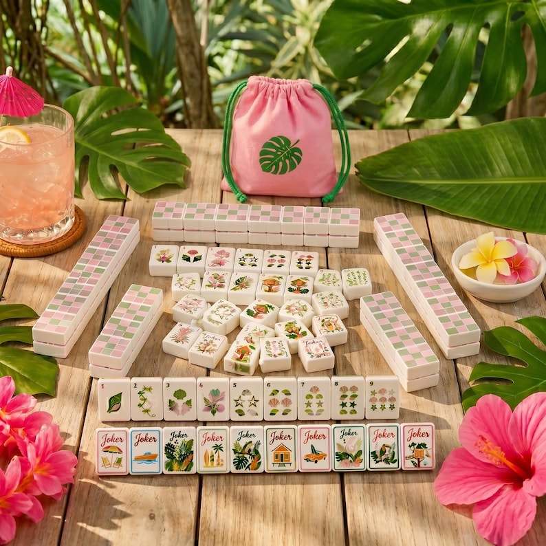 May include: A mahjong set featuring white tiles with colourful botanical and tropical illustrations, including joker tiles with beach-themed graphics. The set includes a pink drawstring storage bag with a green leaf print and four tile racks.