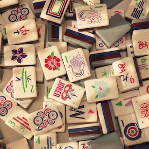 May include: A collection of ivory-colored Mahjong tiles with various colorful designs and symbols. The tiles feature illustrations of flowers, bamboo, and geometric patterns, along with Chinese characters and the word "JOKER". The tiles are arranged in a close-up, overhead shot.