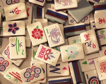 May include: A collection of ivory-colored Mahjong tiles with various colorful designs and symbols. The tiles feature illustrations of flowers, bamboo, and geometric patterns, along with Chinese characters and the word "JOKER". The tiles are arranged in a close-up, overhead shot.