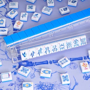 May include: A set of white mahjong tiles with blue backs and blue ink illustrations, including bamboo, character, and dragon designs. A clear plastic rack holds a row of tiles numbered one through nine, while additional loose tiles are scattered.