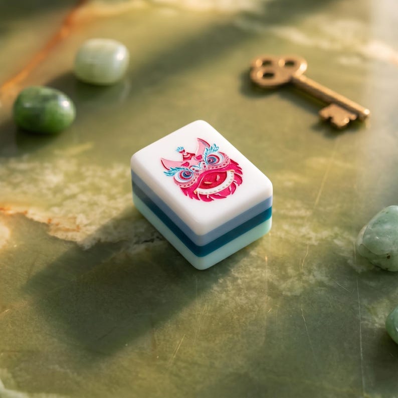 May include: A white and blue Mahjong tile with a colorful lion dance design. The tile is rectangular and stacked with layers of blue and white. A gold key and green stones are in the background.
