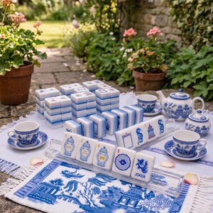 Puede incluir: Un juego de Mahjong completo con fichas azules y blancas, tazas de t&eacute;, una tetera y un azucarero. El juego est&aacute; dispuesto sobre una alfombra estampada en blanco y azul con flecos. La escena est&aacute; ambientada al aire libre con flores al fondo.