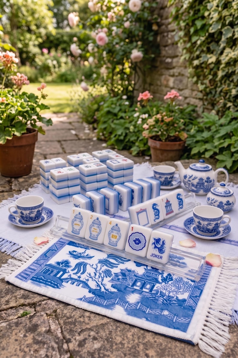 Puede incluir: Un juego de Mahjong completo con fichas azules y blancas, tazas de t&eacute;, una tetera y un azucarero. El juego est&aacute; dispuesto sobre una alfombra estampada en blanco y azul con flecos. La escena est&aacute; ambientada al aire libre con flores al fondo.
