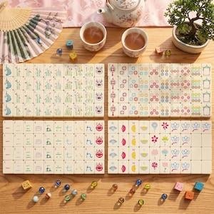 May include: A tabletop arrangement featuring a Mahjong set with ivory tiles, colorful illustrations, and Chinese characters. The scene includes tea cups, a teapot, dice, and decorative marbles, suggesting a game setting.