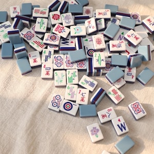 May include: A collection of Mahjong tiles scattered on a light-colored surface. The tiles feature various symbols, numbers, and characters in red, green, and blue, with a light blue border. Some tiles have floral and bamboo designs.