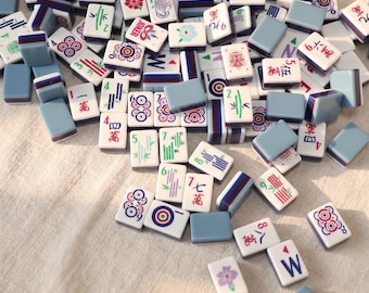 May include: A collection of Mahjong tiles scattered on a light-colored surface. The tiles feature various symbols, numbers, and characters in red, green, and blue, with a light blue border. Some tiles have floral and bamboo designs.