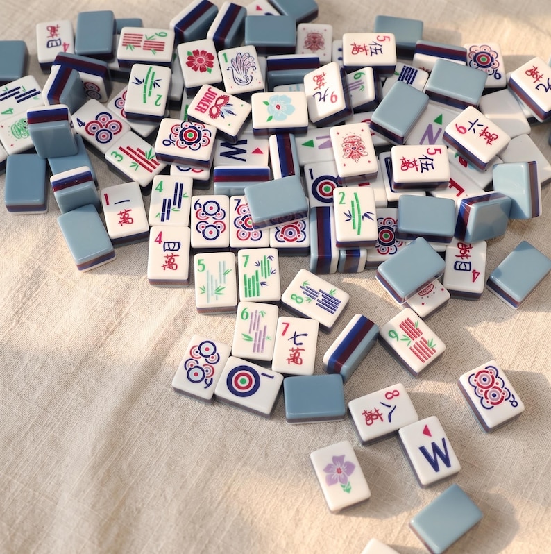 May include: A collection of Mahjong tiles scattered on a light-colored surface. The tiles feature various symbols, numbers, and characters in red, green, and blue, with a light blue border. Some tiles have floral and bamboo designs.