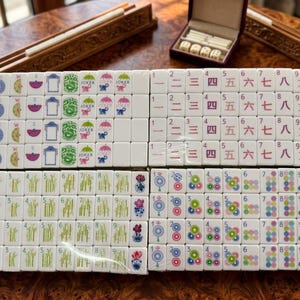 May include: A set of white mahjong tiles arranged in four rectangular blocks. The tiles feature colorful illustrations of fans, compass directions, jokers, monkeys, Chinese numerals, bamboo stalks, circular patterns, and floral motifs in various colors.