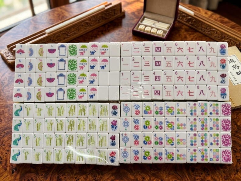 May include: A set of white mahjong tiles arranged in four rectangular blocks. The tiles feature colorful illustrations of fans, compass directions, jokers, monkeys, Chinese numerals, bamboo stalks, circular patterns, and floral motifs in various colors.