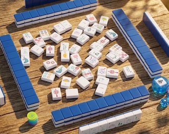 May include: A Mahjong set featuring white tiles with colorful illustrations and Chinese characters, alongside long blue racks for tile organization. The set includes a small stack of circular colored tokens and a translucent blue die on a wooden surface.