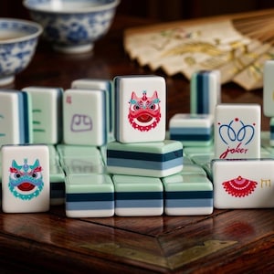 May include: A close-up of a Mahjong set with white tiles featuring colorful illustrations. The tiles have blue, green, and red accents. The set is arranged on a dark wooden surface, with bowls and a fan in the background.