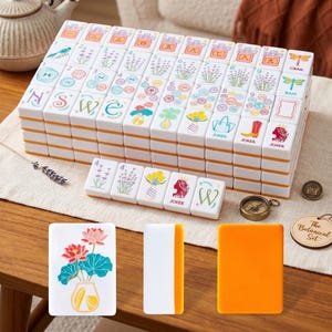 May include: A complete Mahjong set with white tiles featuring colorful floral and botanical illustrations. The tiles are stacked neatly, with orange edges. Additional tiles and accessories are arranged around the main set, including a compass and a wooden tag that says "The Botanical Set."