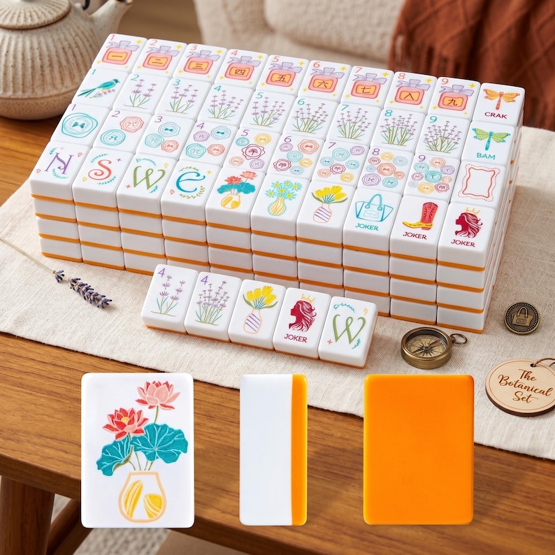May include: A complete Mahjong set with white tiles featuring colorful floral and botanical illustrations. The tiles are stacked neatly, with orange edges. Additional tiles and accessories are arranged around the main set, including a compass and a wooden tag that says "The Botanical Set."