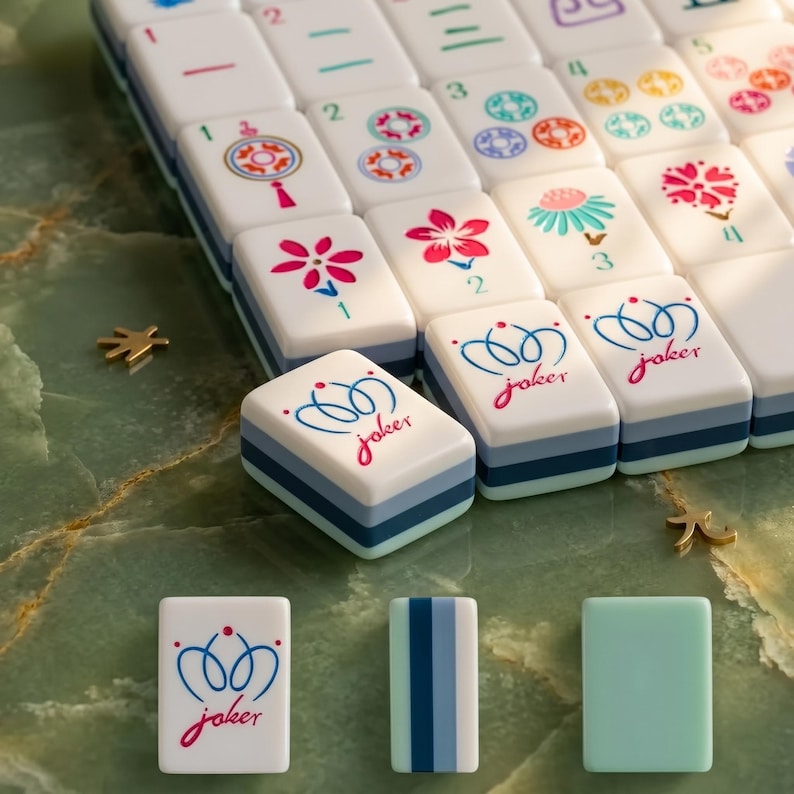 Może przedstawiać: Zestaw białych płytek do gry w Mahjong z kolorowymi wzorami kwiatowymi i liczbowymi. Kilka płytek zawiera słowo "joker" w czerwonym kolorze. Płytki ułożone są na zielonej marmurowej powierzchni, ze złotymi elementami dekoracyjnymi.