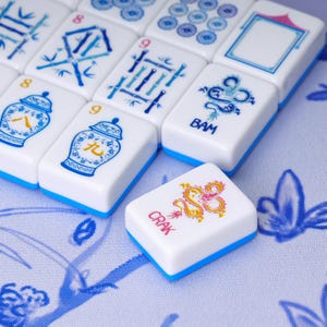 May include: A collection of white Mahjong tiles with blue bases, featuring various blue and red printed designs including bamboo stalks, ginger jars, and dragons. One tile in the foreground displays a red dragon graphic and the text CRAK.