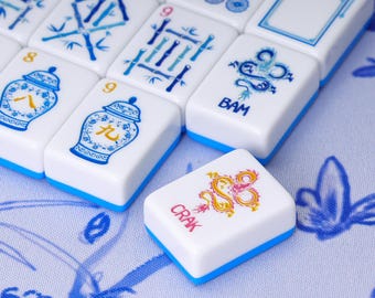 May include: A collection of white Mahjong tiles with blue bases, featuring various blue and red printed designs including bamboo stalks, ginger jars, and dragons. One tile in the foreground displays a red dragon graphic and the text CRAK.