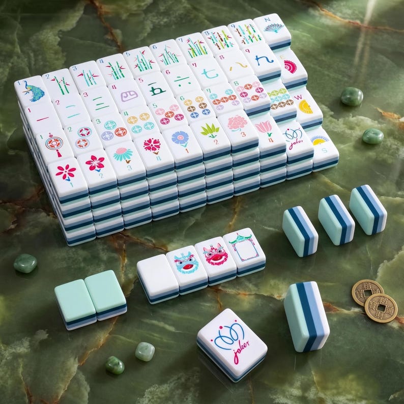 May include: A complete Mahjong set with white tiles featuring various symbols and colorful designs. The set includes dice, tile racks, and decorative coins, all arranged on a green marble surface. The "joker" tile is visible.