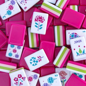 Puede incluir: Una colecci&oacute;n de fichas de Mahjong rosas y blancas. Las fichas presentan varios dise&ntilde;os, incluyendo flores, patrones geom&eacute;tricos y la palabra &laquo;JOKER&raquo;. Las fichas son una mezcla de rosa s&oacute;lido y blanco con acentos coloridos, creando una est&eacute;tica vibrante y juguetona.