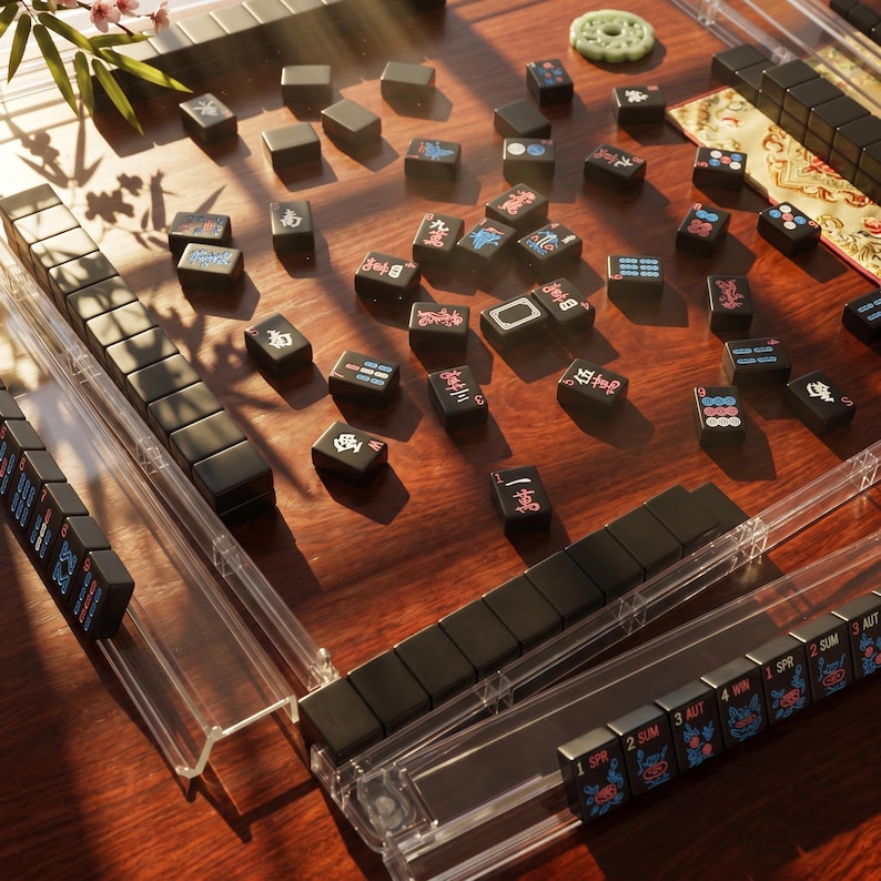 May include: A mahjong set featuring black tiles with colorful Chinese characters and symbols, arranged on a dark wood surface. The set includes clear acrylic tile racks and a round, pale green carved stone piece, with scattered tiles across the board.