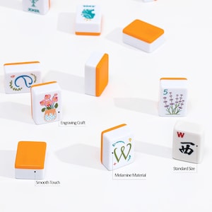 May include: A collection of white and orange Mahjong tiles. Each tile features unique designs, including floral patterns, symbols, and the letter "W". The tiles are labeled "Engraving Craft", "Smooth Touch", "Melamine Material", and "Standard Size".