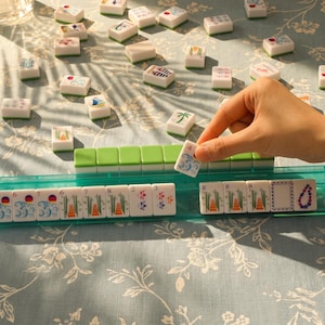 Op de afbeelding: Een Mahjong-spel in uitvoering. De hand houdt een steen met het nummer 4 vast. De stenen zijn wit met diverse kleurrijke symbolen en cijfers. Het spel staat op een lichtblauwe ondergrond met bloemenpatroon.