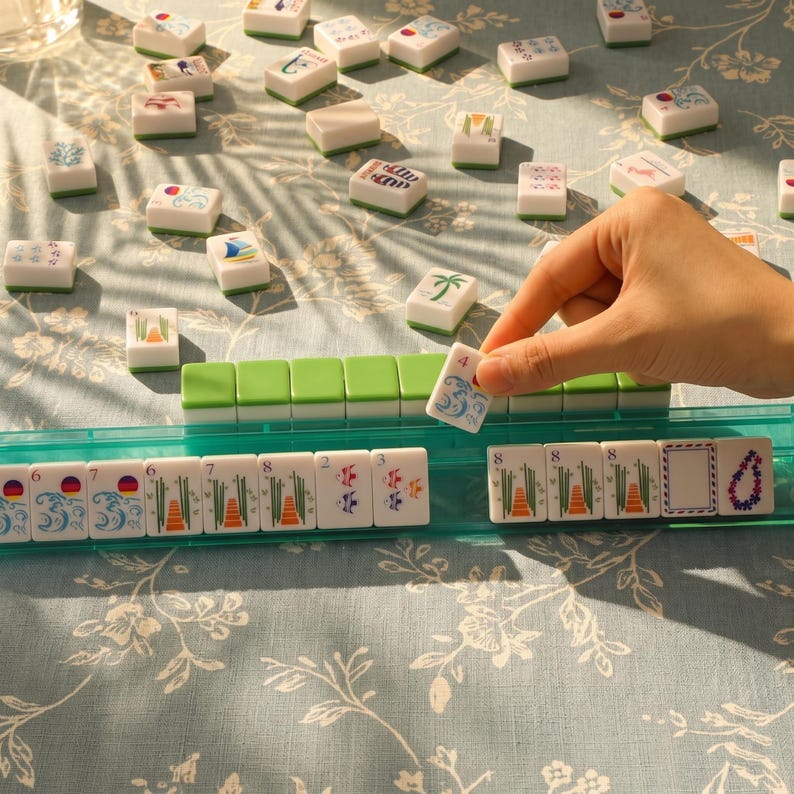 Op de afbeelding: Een Mahjong-spel in uitvoering. De hand houdt een steen met het nummer 4 vast. De stenen zijn wit met diverse kleurrijke symbolen en cijfers. Het spel staat op een lichtblauwe ondergrond met bloemenpatroon.
