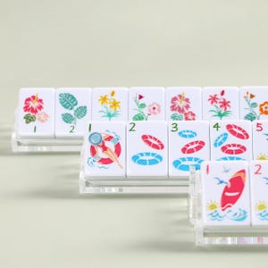 K&ouml;nnte beinhalten: Eine Reihe wei&szlig;er Spielsteine mit farbenfrohen Illustrationen, darunter Blumen, eine Person in einer Schwimmboje und ein Surfbrett. Die Spielsteine sind auf transparenten Acrylst&auml;ndern angeordnet.