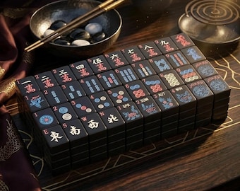 May include: A set of black Mahjong tiles arranged in a rectangular grid. Each tile features engraved characters, numbers, and symbols in red, blue, and white. The tiles display various markings including Chinese characters, directional letters, and Joker labels.