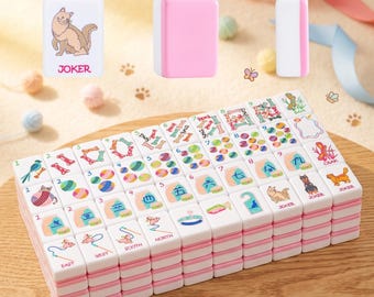 May include: A set of white and pink Mahjong tiles with colorful illustrations and Chinese characters. The tiles feature images of cats, dogs, and other symbols. Some tiles have the word "JOKER" printed on them. The tiles are stacked on a wooden surface.