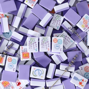 May include: Overhead shot of a collection of lavender and white mahjong tiles. Some tiles feature floral and button designs, while others display numbers. Sprigs of lavender are scattered among the tiles, creating a visually appealing arrangement.