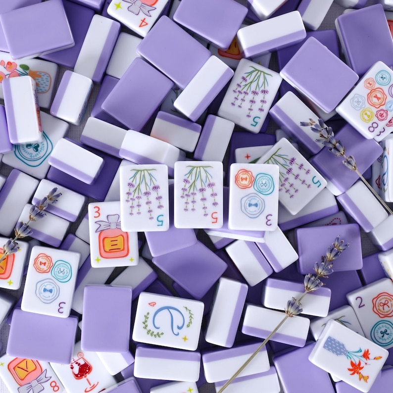 May include: Overhead shot of a collection of lavender and white mahjong tiles. Some tiles feature floral and button designs, while others display numbers. Sprigs of lavender are scattered among the tiles, creating a visually appealing arrangement.