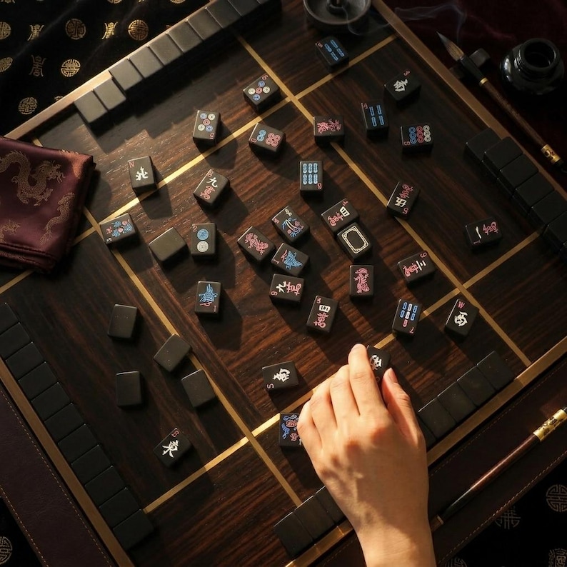 May include: A dark wood mahjong board with a grid pattern features scattered black tiles marked with colorful Chinese characters and symbols. The set includes rows of blank tiles along the edges, a dark fabric cloth with gold patterns, and a writing brush.