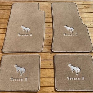 Peut inclure: Un ensemble de quatre tapis de sol en moquette beige pour véhicule. Chaque tapis présente une broderie blanche représentant un cheval cabré et le texte Bronco II. Les deux grands tapis ont un bord profilé, les deux petits sont rectangulaires.