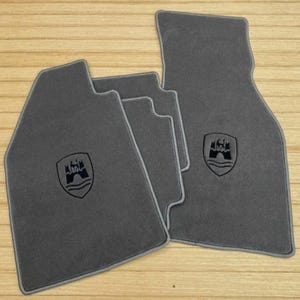 Könnte beinhalten: Ein Set aus vier dunkelgrauen Automatten mit passender grauer Umrandung. Jede Matte ist mit einem schwarzen, gestickten Wappen versehen, das ein Burgtor über wellenförmigen Linien zeigt. Die Matten haben unterschiedliche Formen.