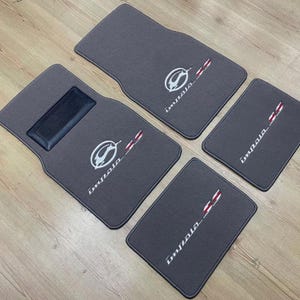 May include: A set of four dark grey automotive floor mats with black trim. Each mat features an embroidered white Impala logo, the text Impala, and a red and white chequered flag emblem. One mat includes a rectangular black heel pad insert.
