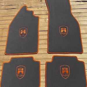 Puede incluir: Un juego de cuatro alfombrillas de coche de color gris oscuro con ribetes naranjas en los bordes. Cada alfombrilla presenta un logotipo de escudo naranja bordado que representa una torre de castillo sobre líneas onduladas, centrado en la superficie.