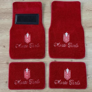 Op de afbeelding: Een set van vier rode tapijt vloermatten voor een voertuig. Elke mat is voorzien van een geborduurd wit Monte Carlo-embleem en tekst. De twee grotere voorste matten bevatten een rechthoekig zwart hielstuk met textuur.