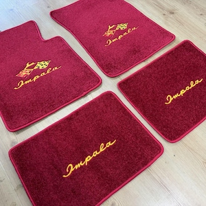 May include: A set of four deep red automotive floor mats with a textured pile surface. Two mats feature an embroidered yellow Impala logo with a checkered flag emblem, while the other two mats display the yellow embroidered text Impala.