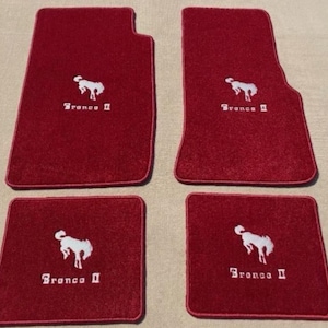 Puede incluir: Un juego de cuatro alfombrillas de coche de color rojo intenso con el logotipo Bronco II bordado en blanco y un gráfico de un caballo encabritado. El juego incluye dos alfombrillas delanteras grandes con esquina recortada y dos traseras.