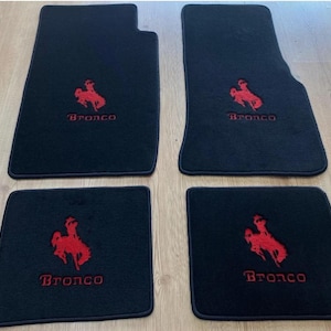 Puede incluir: Un juego de cuatro alfombrillas negras para coche con un logotipo bordado en rojo de un caballo encabritado y la palabra Bronco en letras rojas. Las dos alfombrillas delanteras son más grandes y contorneadas, las traseras son rectangulares.