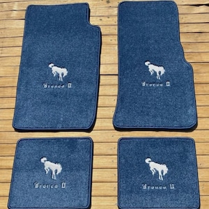 Puede incluir: Un juego de cuatro alfombrillas de coche azul oscuro con un logotipo bordado en blanco de un caballo encabritado y el texto Bronco II. Las dos alfombrillas delanteras son más grandes y contorneadas, mientras que las traseras son rectangulares.