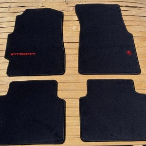 May include: A set of four black automotive floor mats with red trim. The front driver-side mat features the red embroidered text INTEGRA, while the front passenger-side mat displays a small red circular logo. The mats have a carpeted texture.