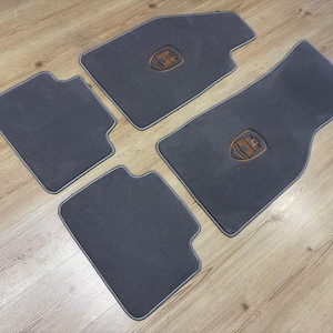 May include: A set of four dark grey automotive floor mats with light grey piping along the edges. Two of the mats feature a centred, shield-shaped emblem in a tan colour with a dark graphic design, while the other two mats are plain.