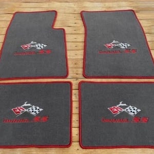 May include: A set of four dark gray automotive floor mats with red trim. Each mat features an embroidered logo with two crossed checkered flags and the red text Impala SS. The mats are rectangular with one mat having a notched corner design.
