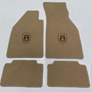 Puede incluir: Un juego de cuatro alfombrillas de coche color canela con superficie de moqueta texturizada. Las dos alfombrillas delanteras más grandes tienen un emblema de escudo bordado en marrón oscuro con una puerta de castillo y ondas de agua.