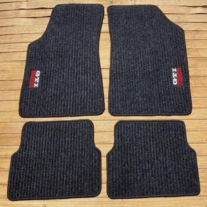 Könnte beinhalten: Ein Set aus vier dunkelgrauen Auto-Fußmatten mit vertikaler Rippenstruktur. Jede Matte verfügt über ein rechteckiges, rot-weiß besticktes GTI-Logo. Die beiden größeren vorderen Matten haben eine abgeschrägte obere Ecke, die hinteren sind rechteckig.
