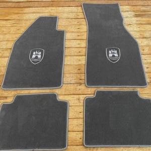 Puede incluir: Un juego de cuatro alfombrillas de coche de color gris oscuro con ribetes de color gris claro en los bordes. Dos de las alfombrillas presentan un logotipo de escudo bordado en blanco que representa un castillo sobre una línea ondulada.
